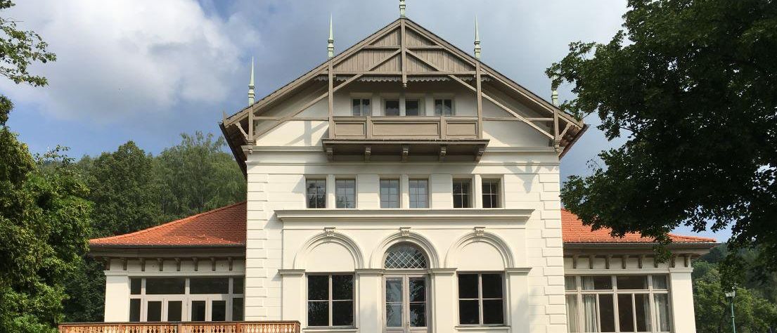 Ein großes weißes Gebäude mit einem braunen Dach und einem hölzernen Balkon. Es hat viele Fenster und einen kleinen Vorbau mit Topfpflanzen. Es steht auf einem grünen Hügel.