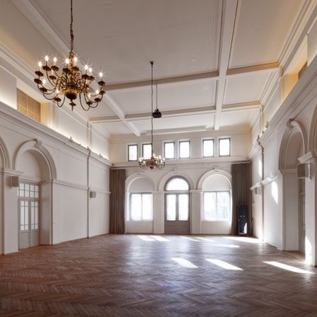 Ein leerer Raum mit hohen Decken, Holzböden und zwei Kronleuchtern. Es gibt bogenförmige Fenster und Türen, und die Wände sind weiß.