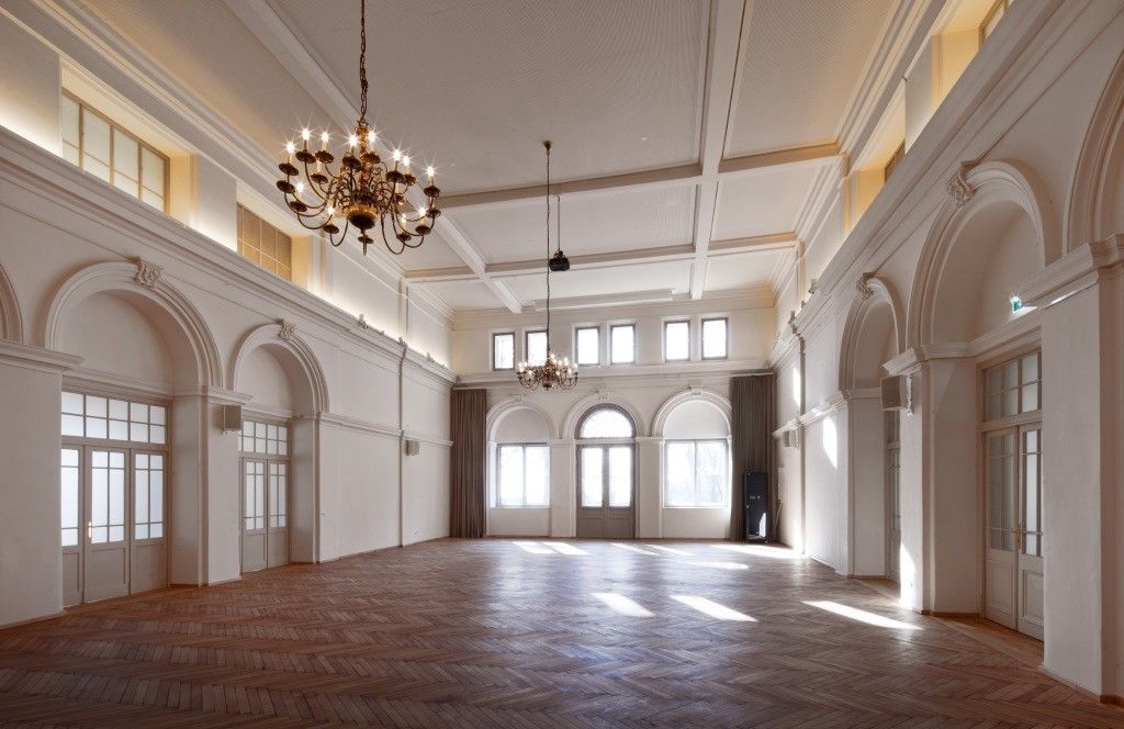 Ein leerer Raum mit hohen Decken, Holzböden und zwei Kronleuchtern. Es gibt bogenförmige Fenster und Türen, und die Wände sind weiß.