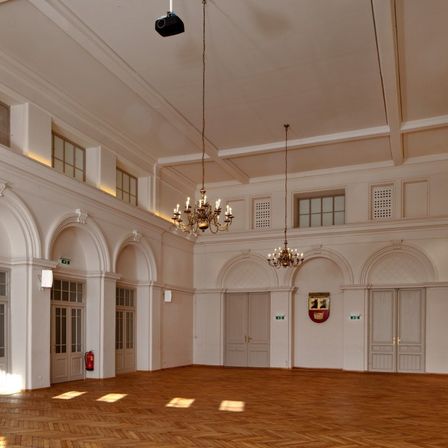 Ein leerer Raum mit weißen Wänden, Holzböden und gewölbten Türen. Zwei Kronleuchter hängen von der Decke. Ein Beamer ist über dem Raum montiert.
