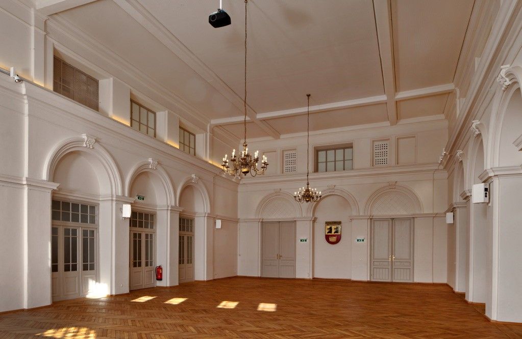 Ein leerer Raum mit weißen Wänden, Holzböden und gewölbten Türen. Zwei Kronleuchter hängen von der Decke. Ein Beamer ist über dem Raum montiert.