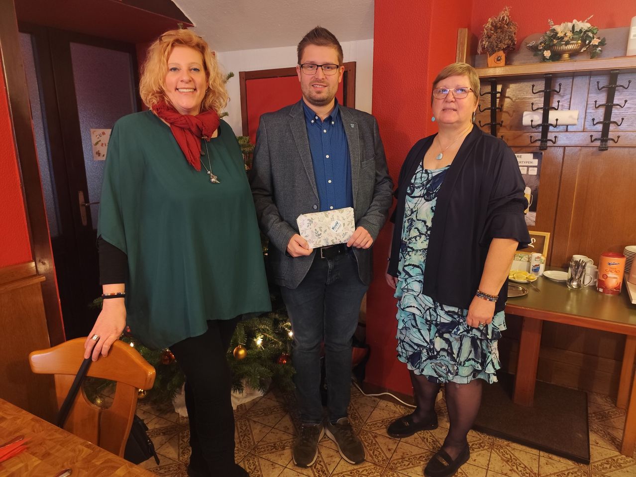 Drei Personen stehen neben einem Weihnachtsbaum, der Mann hält ein Geschenk. Eine Frau links trägt ein grünes Oberteil und einen roten Schal, und eine andere Frau rechts trägt ein blaues Kleid.