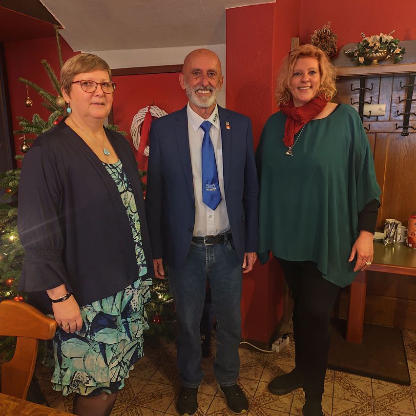 Drei Menschen stehen nebeneinander vor einem Weihnachtsbaum und tragen formelle Kleidung. Der Mann hat eine Anstecknadel an seiner Jacke.