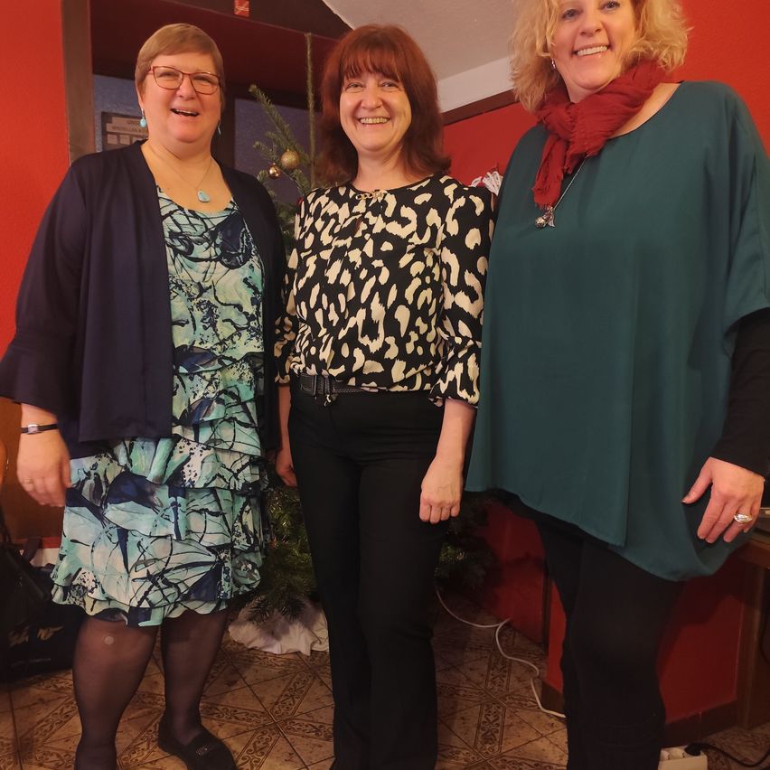 Drei Frauen in festlicher Kleidung stehen nah beieinander und lächeln, mit einem Weihnachtsbaum im Hintergrund.