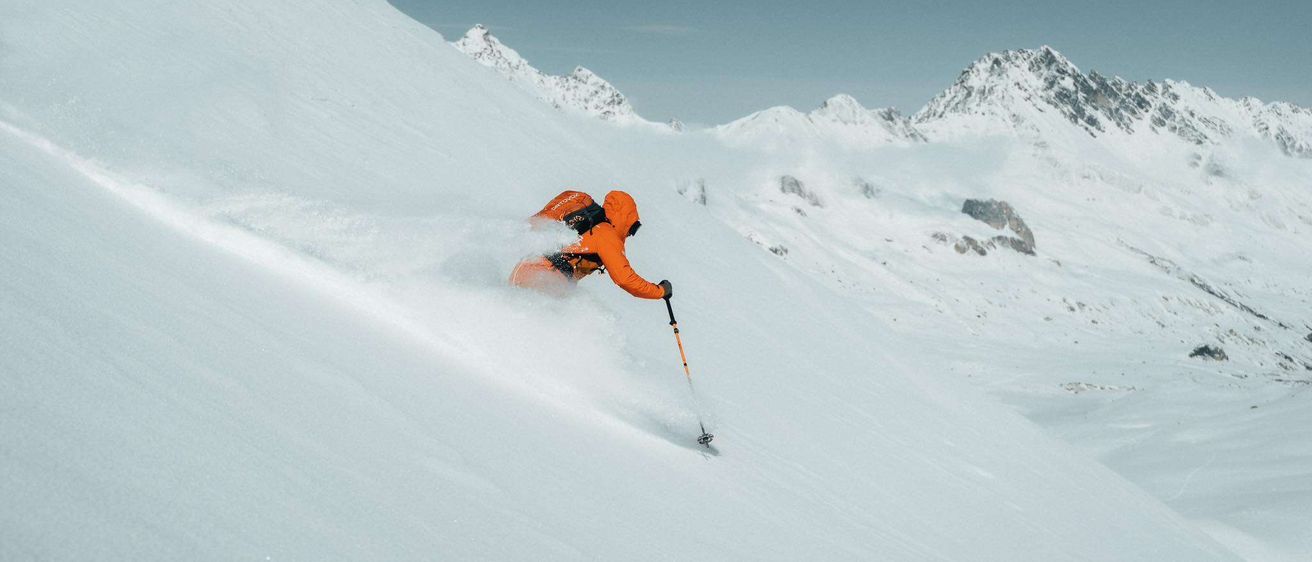 Eine Person, die einen verschneiten Berg hinunterskifahrert, trägt einen orangefarbenen Anorak und einen Rucksack. Schnee fliegt um sie herum.