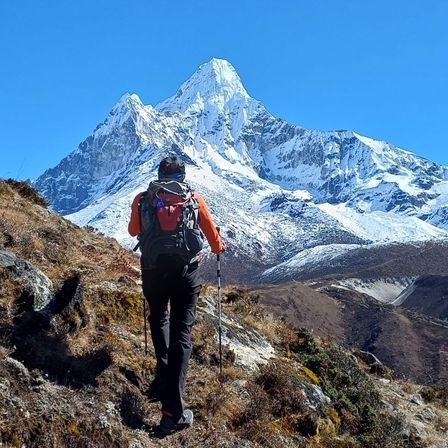 Ein Wanderer mit Rucksack erklimmt einen Berghang unter blauem Himmel. Im Hintergrund sind schneebedeckte Gipfel zu sehen.