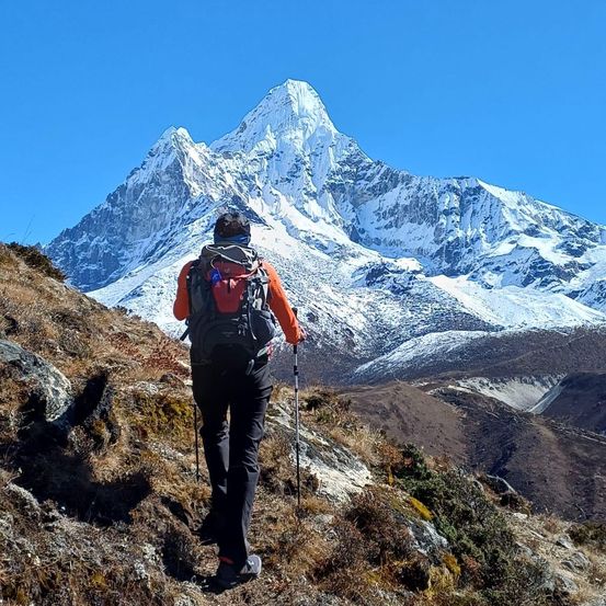Ein Wanderer mit Rucksack erklimmt einen Berghang unter blauem Himmel. Im Hintergrund sind schneebedeckte Gipfel zu sehen.