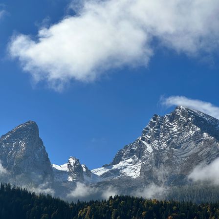 Ein klarer blauer Himmel mit einer Wolke über verschneiten Berggipfeln, umgeben von dichtem grünem Wald.