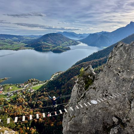 Eine Person in einem blauen Outfit und Helm hängt von einer Seilbrücke über einem See, umgeben von Bergen und Bäumen.