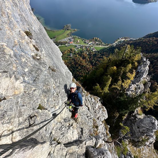 Ein Kletterer erklimmt eine steile Felswand, während er einen Helm und eine Sonnenbrille trägt. Unten gibt es eine friedliche Landschaft mit einem See und grünen Feldern.