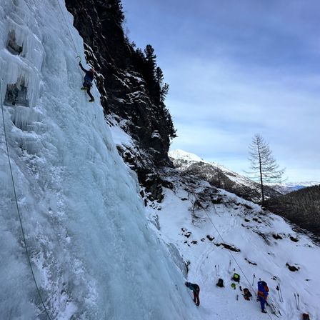 Eine Gruppe von Kletterern erklimmt einen gefrorenen Wasserfall in den verschneiten Bergen. Die Kletterer sind mit Seilen und Eispickeln ausgestattet.