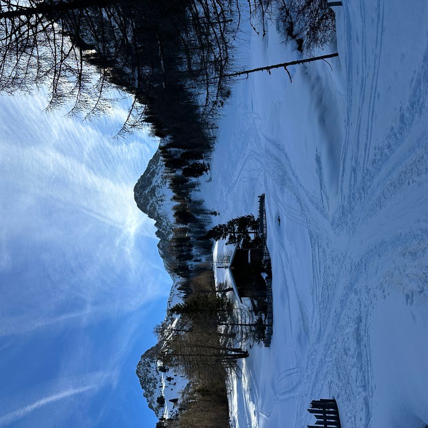 Eine Luftaufnahme eines verschneiten Berges mit einem Gebäude und Bäumen. Der Berg ist schneebedeckt und der Himmel ist blau mit einigen Wolken.