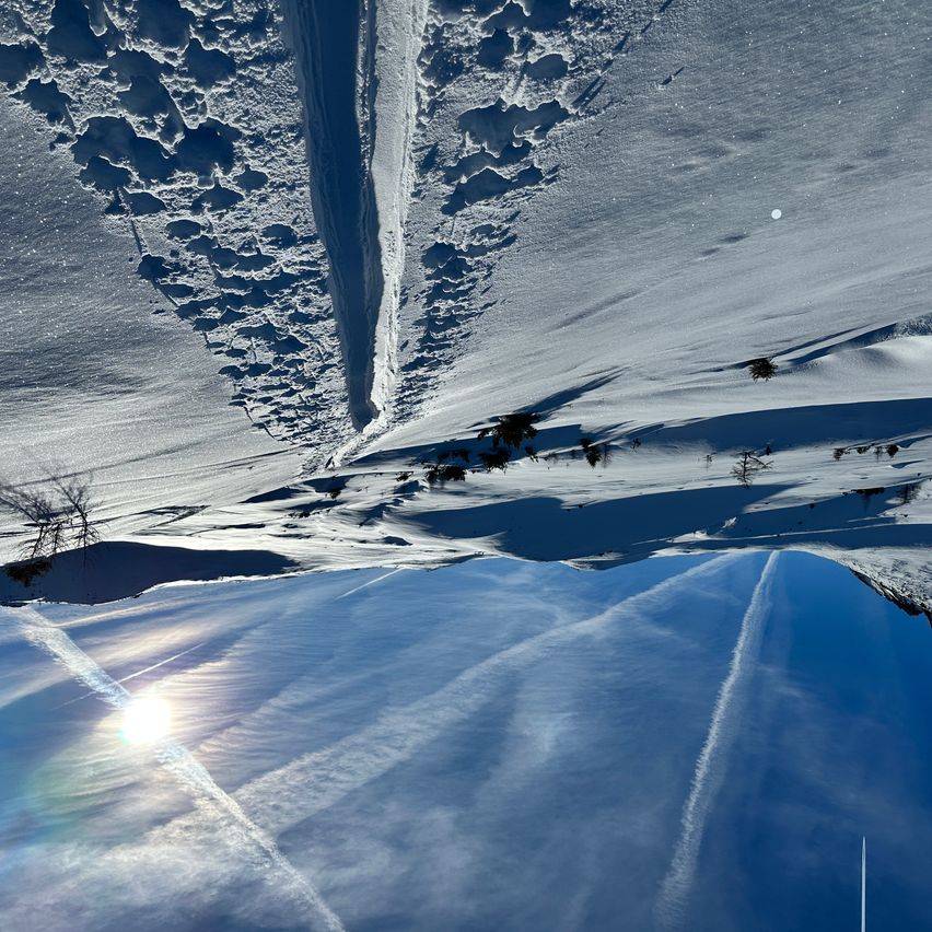 Luftaufnahme von verschneiten Bergen mit tiefen Furchen und Spuren, einer hellen Sonne, die einen Regenbogen am Himmel wirft, und Kondensstreifen von Flugzeugen.