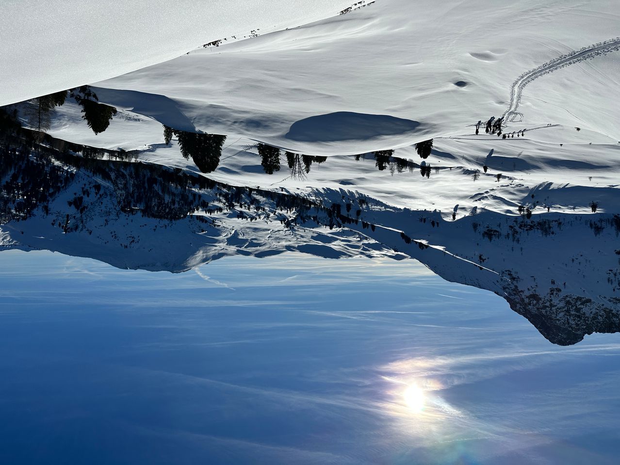 Eine verschneite Berglandschaft mit einem hellen Sonnenschein, der auf dem Schnee reflektiert und einen strahlenden Effekt erzeugt, und einer Gruppe von Skifahrern auf einer Loipe.