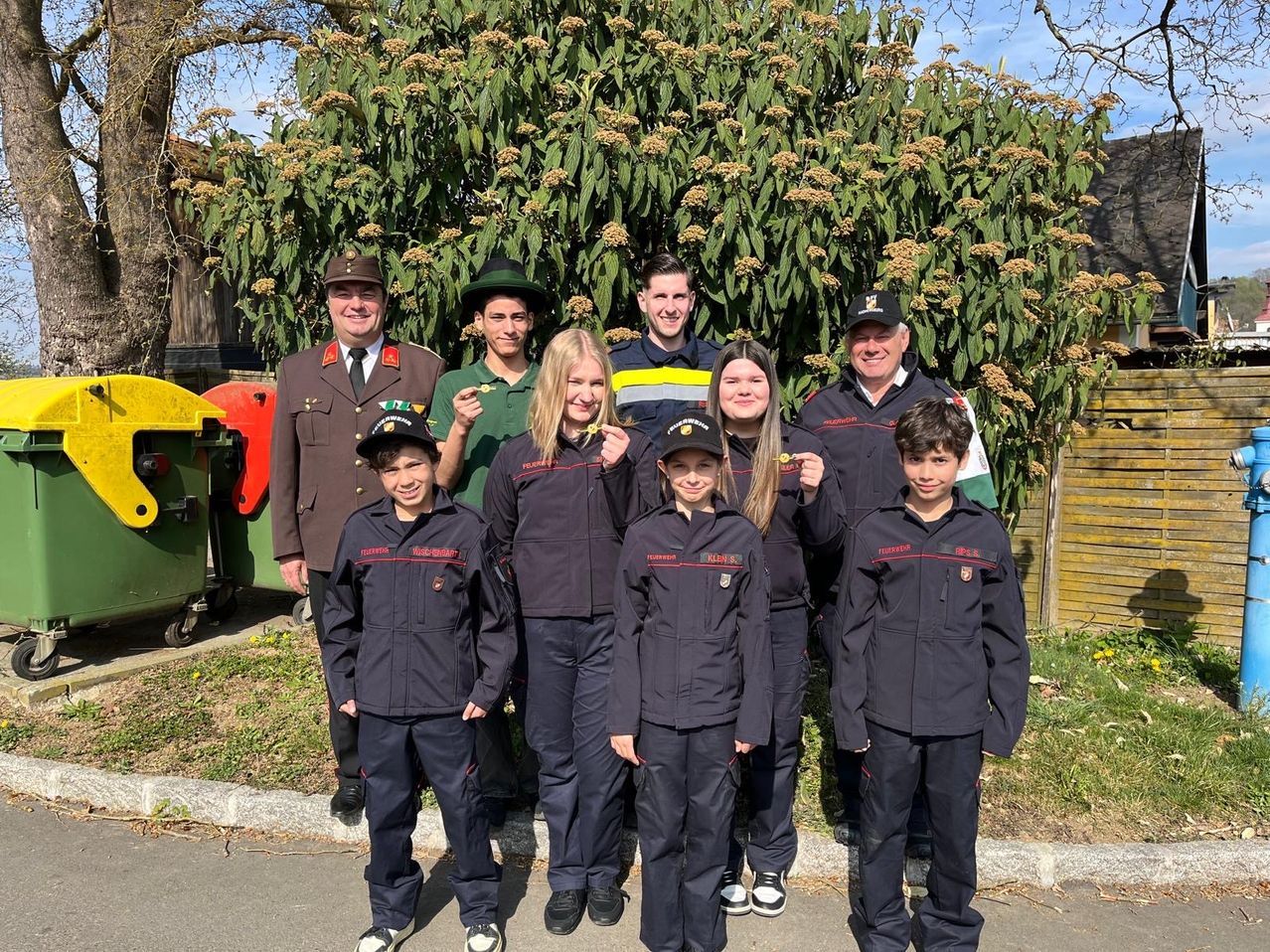 A group of young people in uniforms with hats and badges stand with a smiling adult man in military uniform. Behind them is a bush with many flowers.