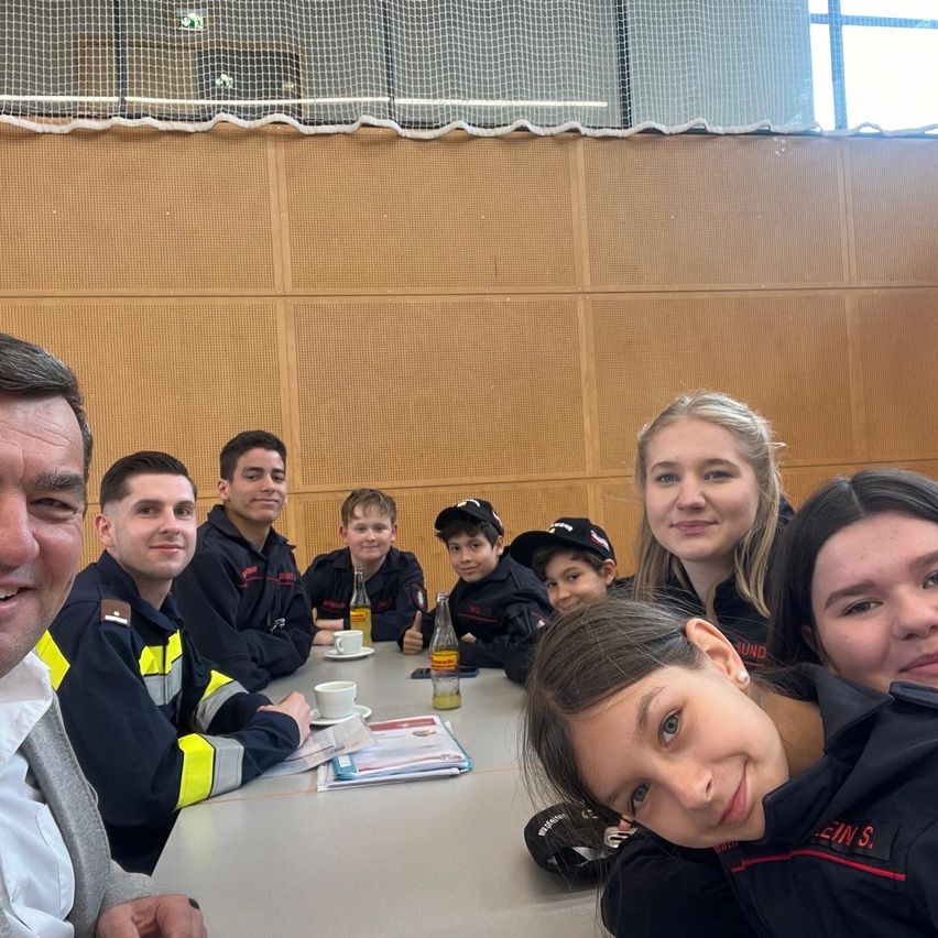 A group of people, possibly firefighters, are seated at a table indoors, posing for a photo. They wear uniforms with the word 'Feuerwehr' and are smiling. There are cups, plates, and a bottle on the table. The wall is made of wood.
