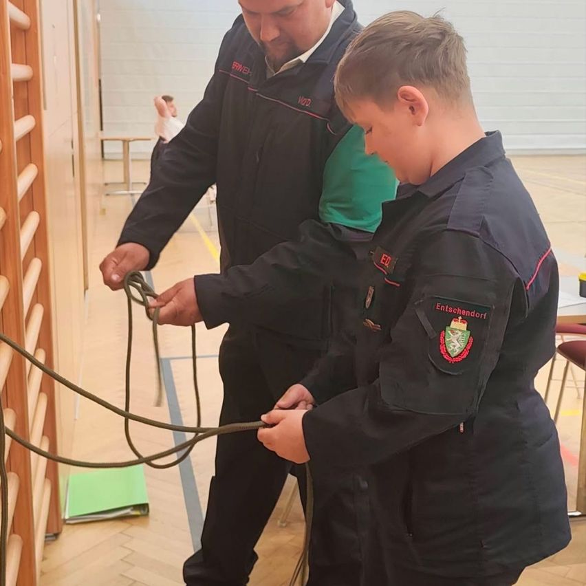 A man and a boy in uniforms are tying a rope together in a room. A woman stands behind them.