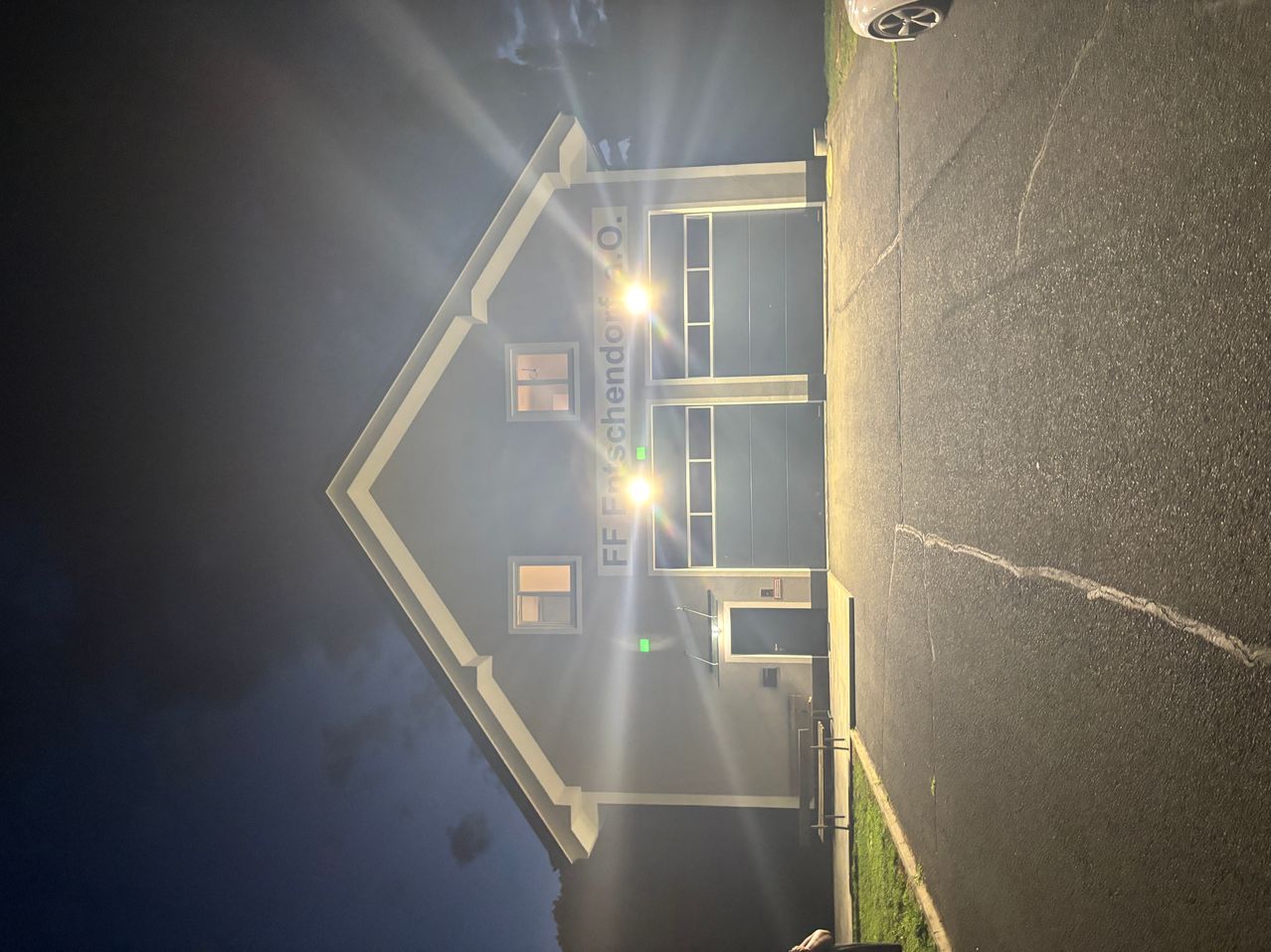 Ein Gebäude mit dem Namen FF Fischendorf O. ist nachts mit zwei hellen Lichtern beleuchtet. Das Gebäude hat eine dreieckige Form mit zwei Fenstern und einer Tür. Das Gebäude ist von einem Betonweg und Gras umgeben.
