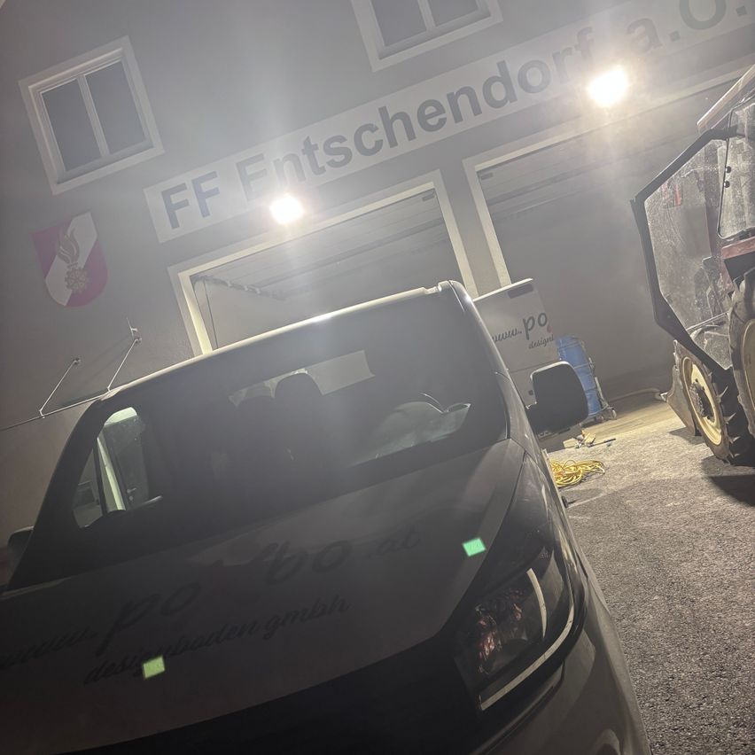 A car is parked in front of a building with the name FF Entschendorf. The headlights are on, and the car's windshield is foggy.
