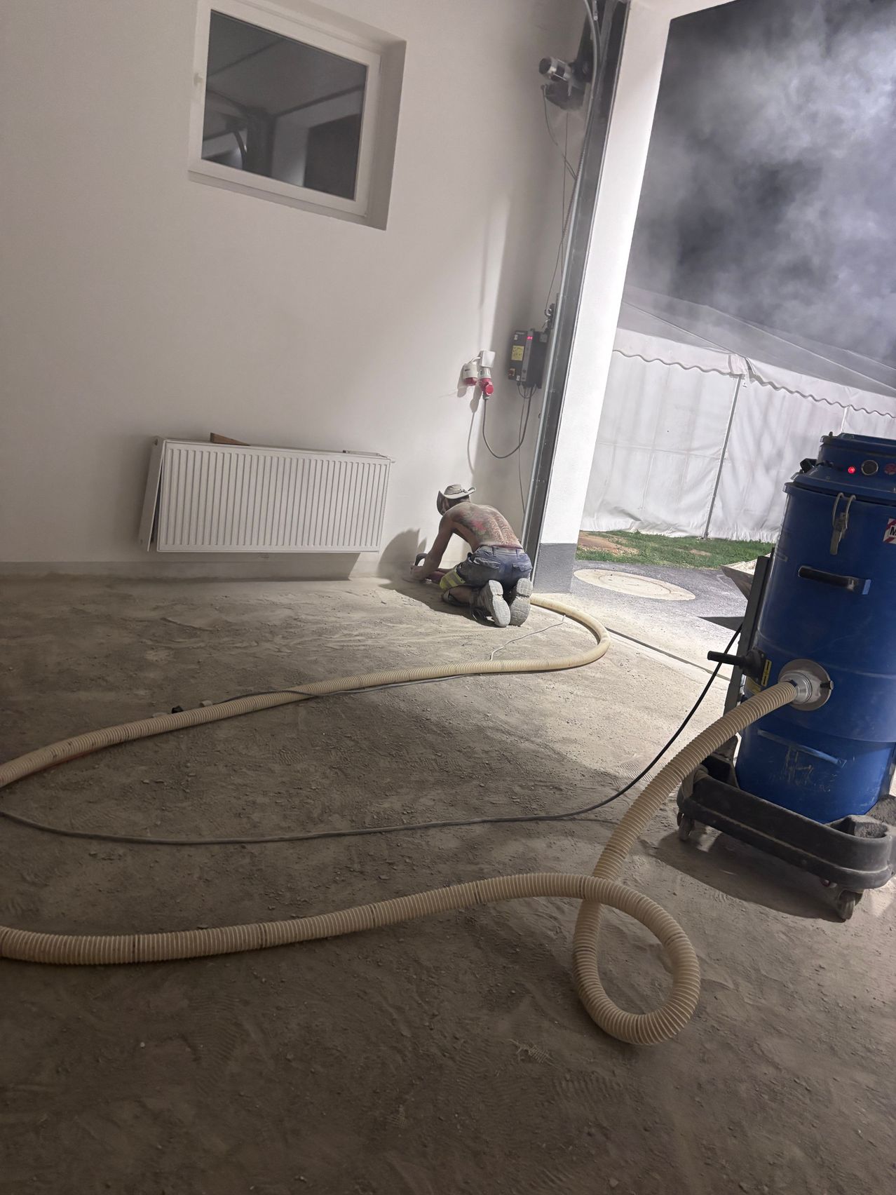 A man is kneeling on the floor, using a grinder. A large hose is connected to a blue vacuum. The room has a white wall and a white radiator.
