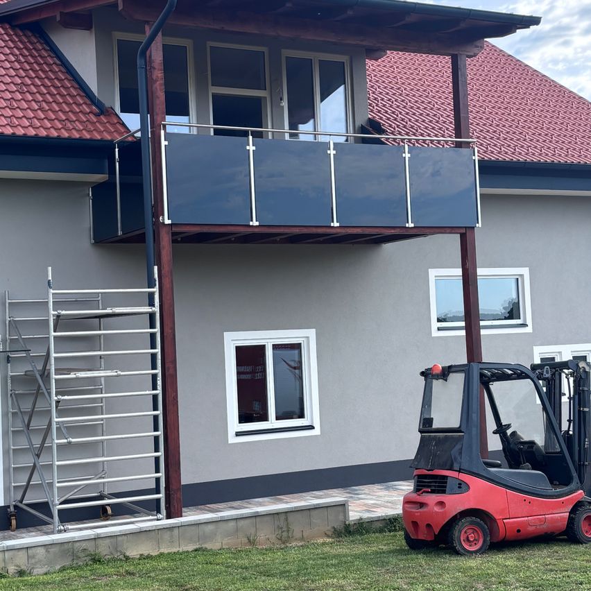 A house with a red roof and a balcony with a glass railing is under construction. A red forklift is parked in front.