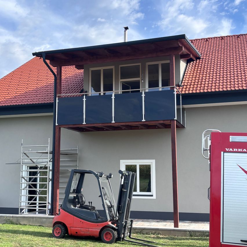A red forklift is parked in front of a house with a balcony, under a partly cloudy sky.