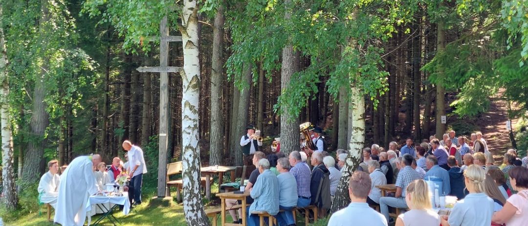 Eine Gruppe von Menschen versammelt sich in einer Waldlichtung für eine musikalische Veranstaltung. Eine Person steht an einem Tisch, und Musiker spielen Instrumente. Ein Priester bereitet sich in der Nähe vor. Die Menge sitzt auf Bänken und Stühlen.