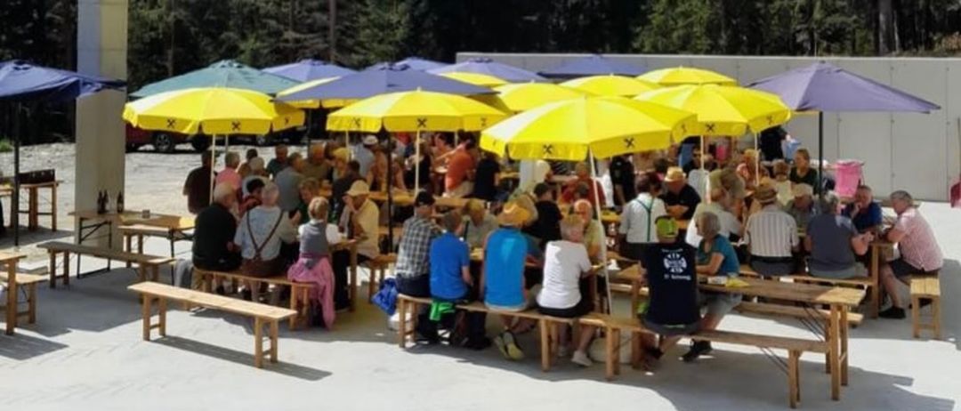 Eine Gruppe von Menschen sitzt an Picknicktischen unter gelben Sonnenschirmen mit Tannen im Hintergrund.