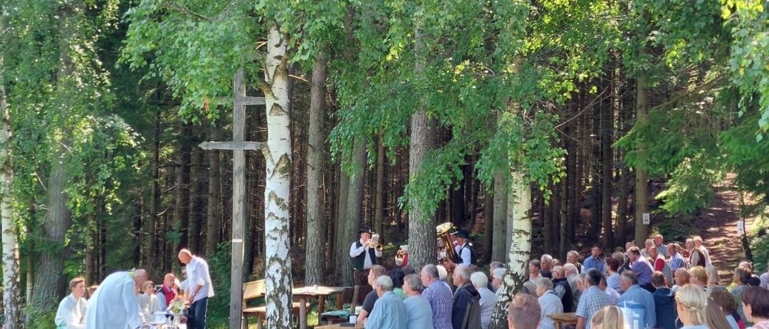 A large group gathers in a forest clearing for an event. Some are seated while others stand. Two men play brass instruments, and a man officiates a ceremony. Trees surround the area, with a cross on a pole nearby.