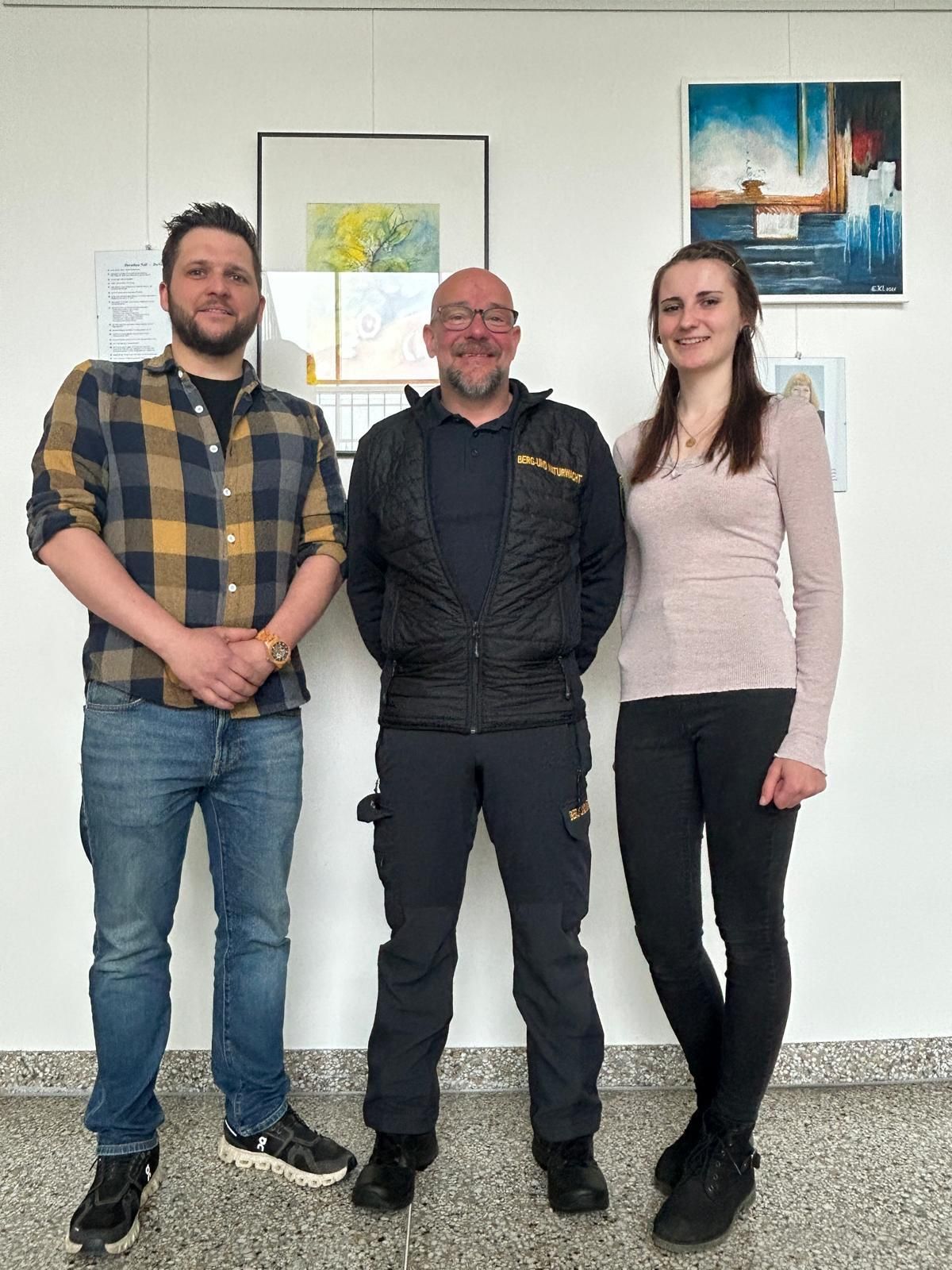 Drei Personen stehen gemeinsam vor einer weißen Wand mit Gemälden. Der Mann links trägt ein kariertes Hemd und der Mann in der Mitte ein schwarzes Jacket. Die Frau rechts trägt ein helles rosa Oberteil und schwarze Hosen.