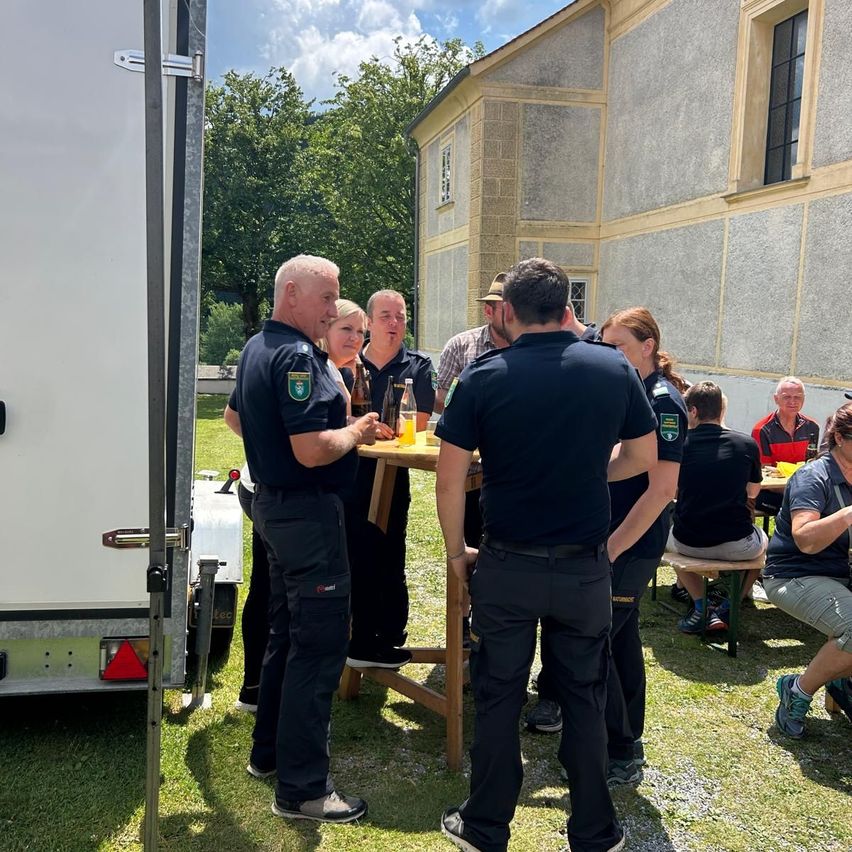 Eine Gruppe uniformierter Personen, möglicherweise Feuerwehrleute, versammelt sich um einen Tisch mit Getränken in einer Außenumgebung nahe eines Gebäudes.