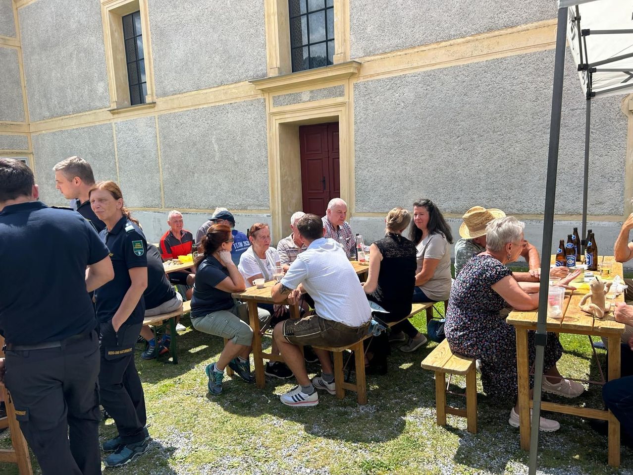 Menschen sitzen an Holztischen vor einem Gebäude in Österreich, bei einer Veranstaltung.