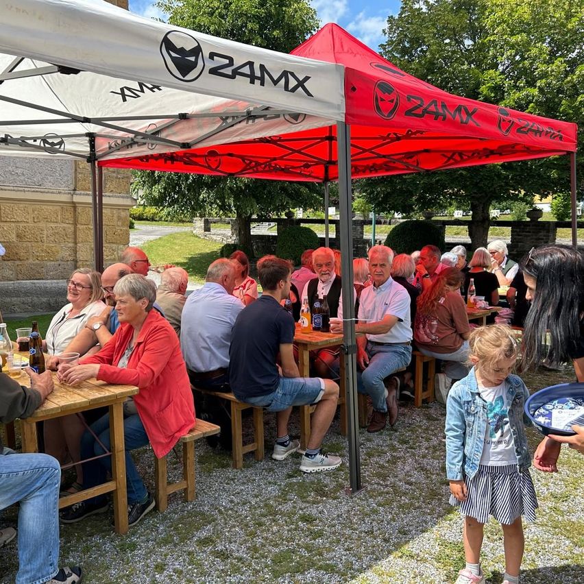 Eine Gruppe von Menschen sitzt unter einem roten Zelt mit dem Wort Zamx darauf. Sie essen und trinken. Ein kleines Mädchen steht vor einem Tisch mit einem Tablett.