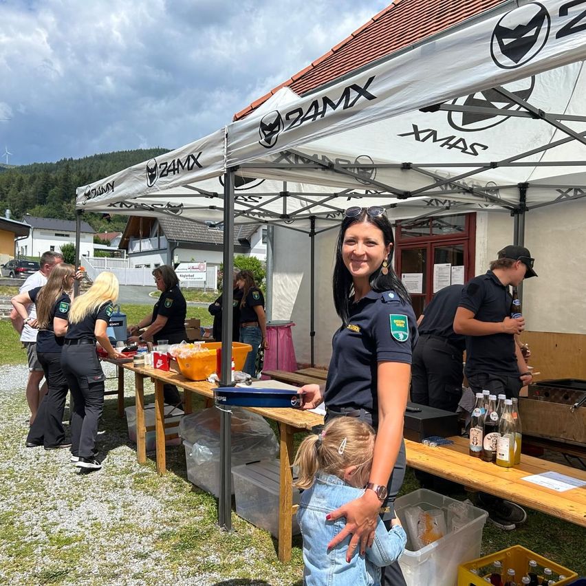 Eine Frau in Uniform steht unter einem Zelt mit einem lächelnden Mädchen, während andere Essen auf Tischen zubereiten. Ein Gebäude und Berge sind im Hintergrund.