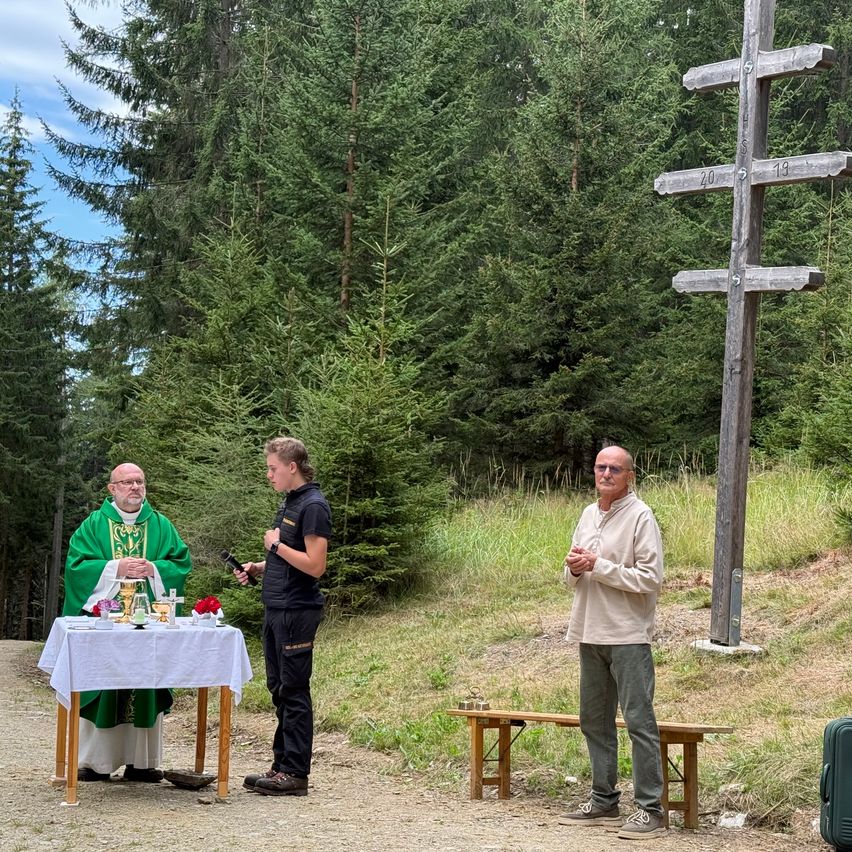 Ein Priester in einem grünen Gewand steht hinter einem Tisch mit einem Mikrofon, während ein Mann in einer schwarzen Jacke neben ihm steht. Ein anderer Mann in einer weißen Jacke steht rechts. Ein hölzernes Kreuz steht hinter ihnen.