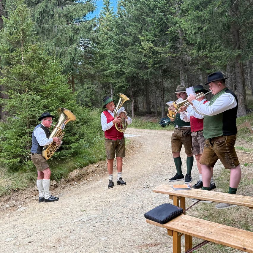 Eine Gruppe von Musikern in traditioneller bayerischer Kleidung spielt Blechblasinstrumente auf einem unbefestigten Weg, umgeben von Bäumen.