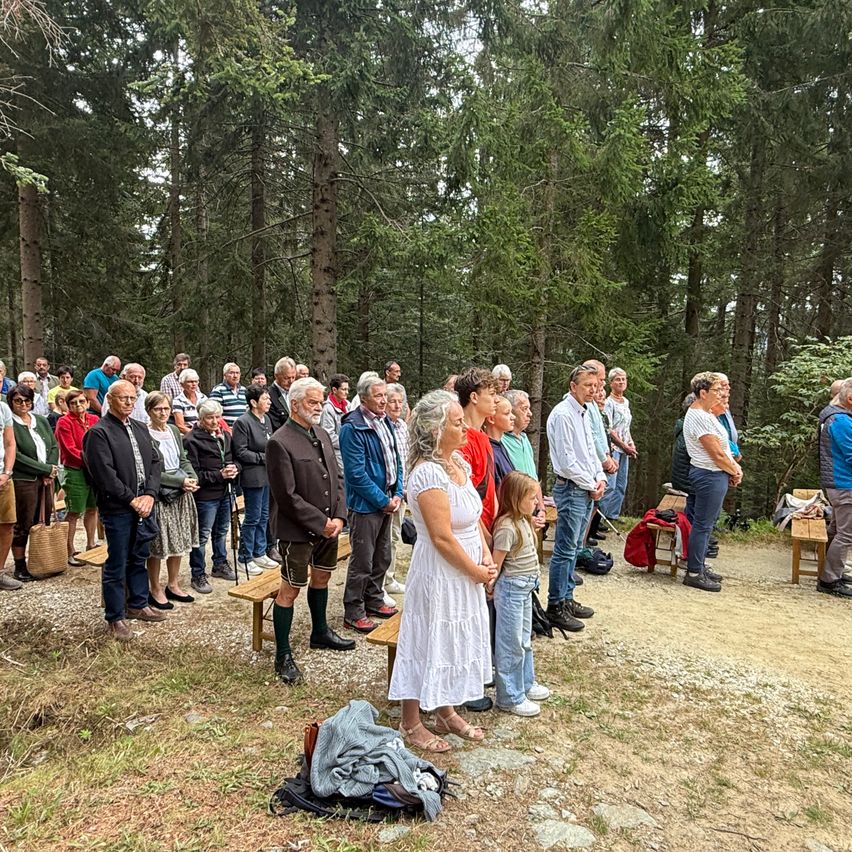 Eine Gruppe von Menschen versammelt sich in einer Waldlichtung, einige stehen, andere sitzen auf Bänken. Sie scheinen an einer Zeremonie oder Versammlung teilzunehmen, mit einer Frau in einem weißen Kleid und einem jungen Mädchen in der Mitte.