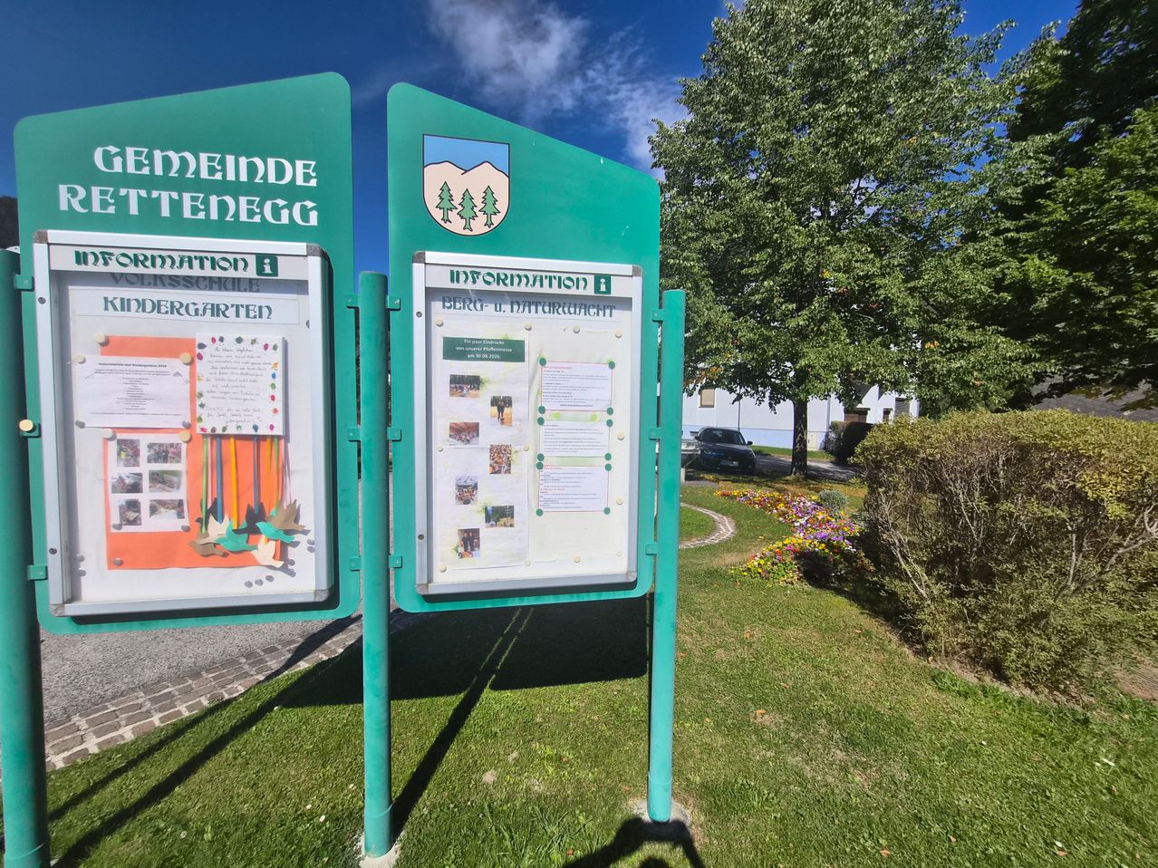 A green informational signboard stands on a grassy lawn with a tree and flowers in the background. The signboard has a logo and text in German.