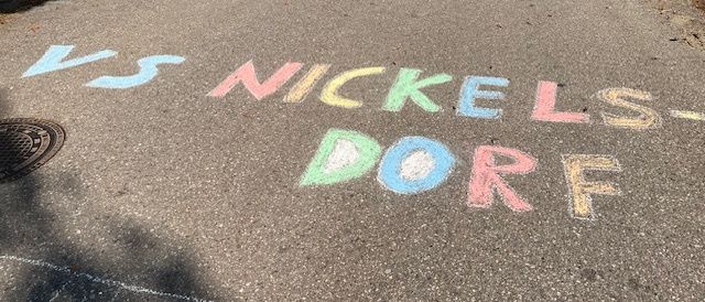 Ein Bürgersteig mit einer bunten Kreidezeichnung von 'VS NICKELSDORF' auf dem Bürgersteig. Ein Kanaldeckel ist in der Nähe. Mehrere Menschen gehen auf dem Bürgersteig.