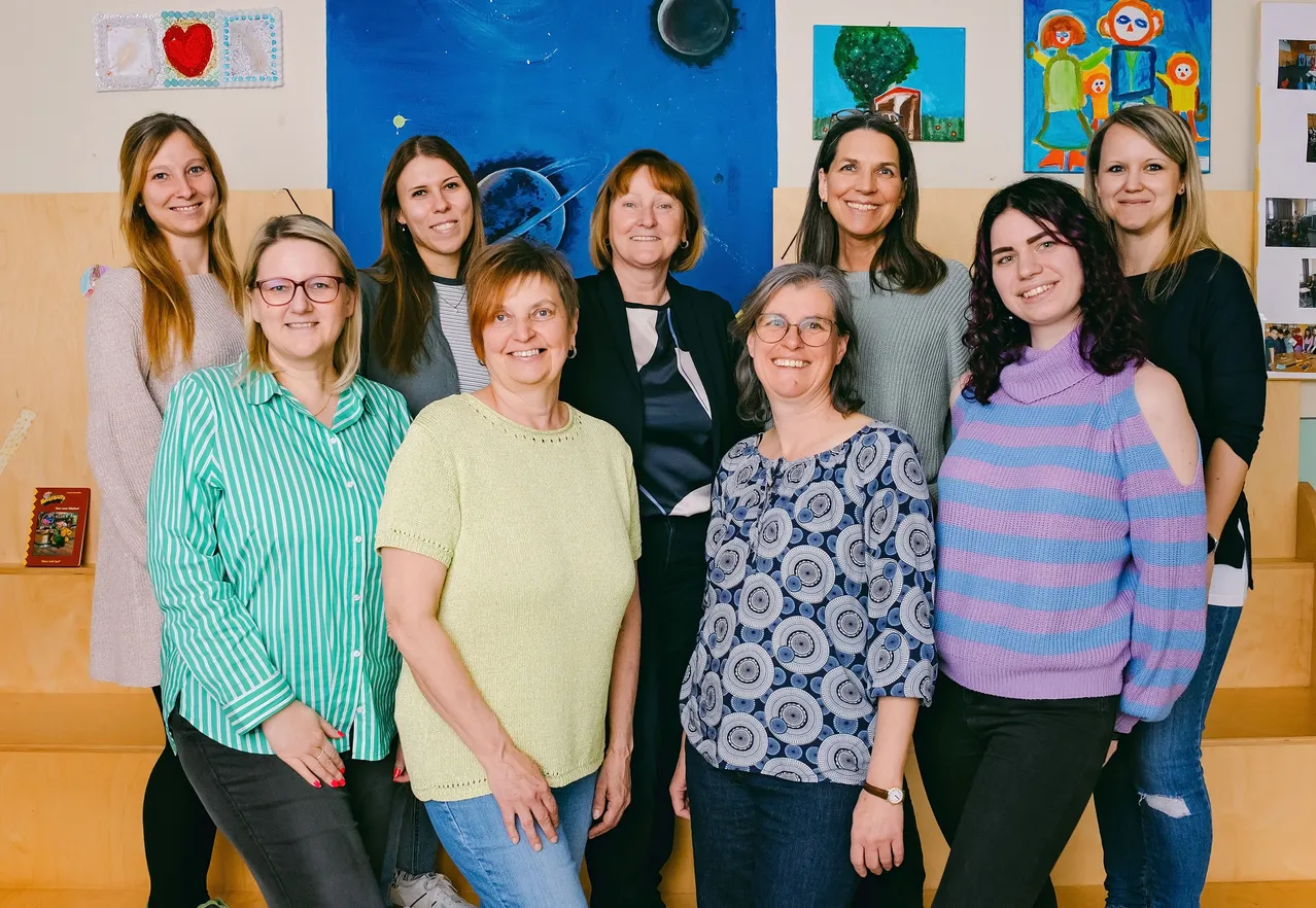Eine Gruppe erwachsener Frauen in verschiedenen Outfits posiert für ein Foto vor einer Wand mit farbenfrohen Gemälden.