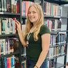 Eine junge Frau mit blonden Haaren steht in einer Bibliothek, berührt ein Regal voller Bücher und trägt ein grünes T-Shirt und Jeansshorts.