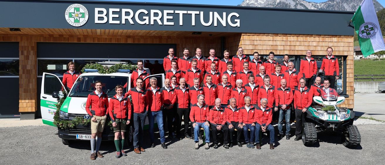 Eine Gruppe von Menschen in roten Jacken posiert vor einem Gebäude mit einem verschneiten Berg im Hintergrund.