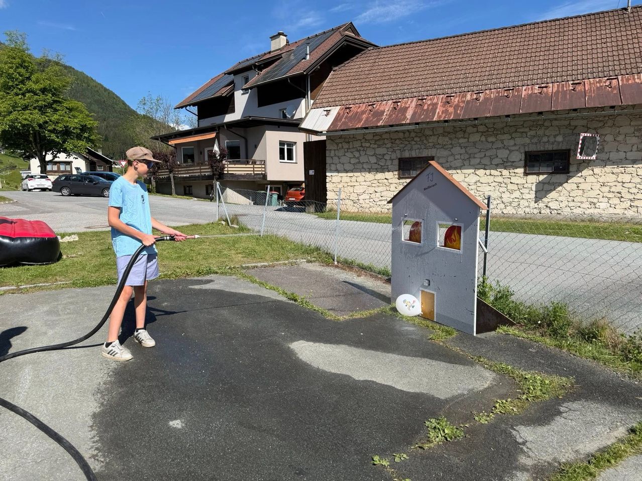 Ein junger Junge benutzt einen Schlauch, um ein kleines hausartiges Gebäude vor einem Steingebäude mit Solarmodulen zu bewässern.