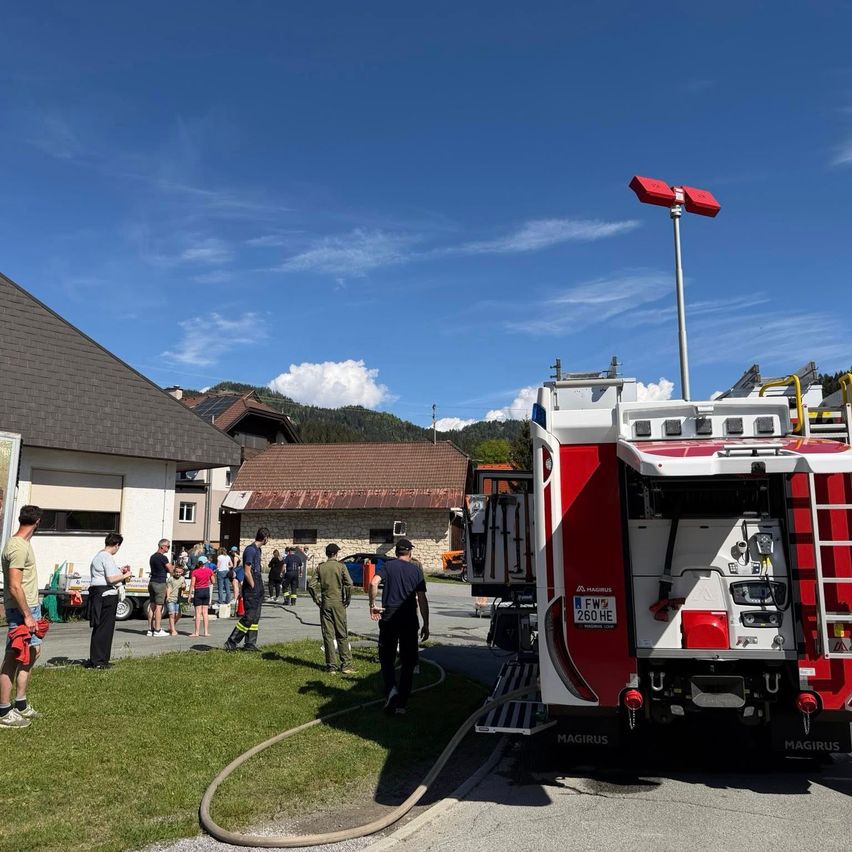 Ein roter Feuerwehrwagen ist neben einem Gebäude geparkt. Menschen stehen herum, einige tragen Uniformen. Der Himmel ist klar und blau.