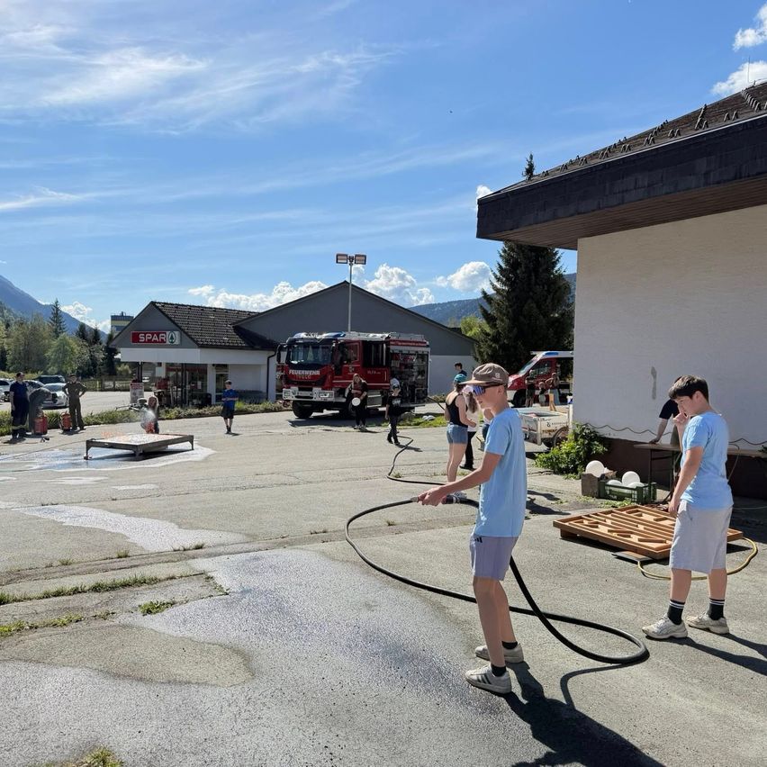 Zwei Jungen säubern eine Straße mit einem Schlauch. Dahinter steht ein Feuerwehrauto, ein Gebäude und ein Geschäft. Eine Gruppe von Menschen ist in der Nähe.