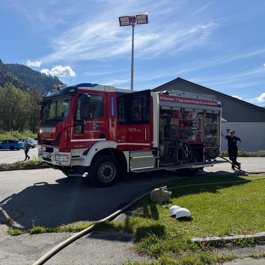 Ein roter Feuerwehrwagen ist auf einer Straße geparkt, mit einer angeschlossenen Feuerwehrschlauch. Ein Mann steht neben ihm. Hinter dem LKW sind mehrere geparkte Autos und ein Gebäude.
