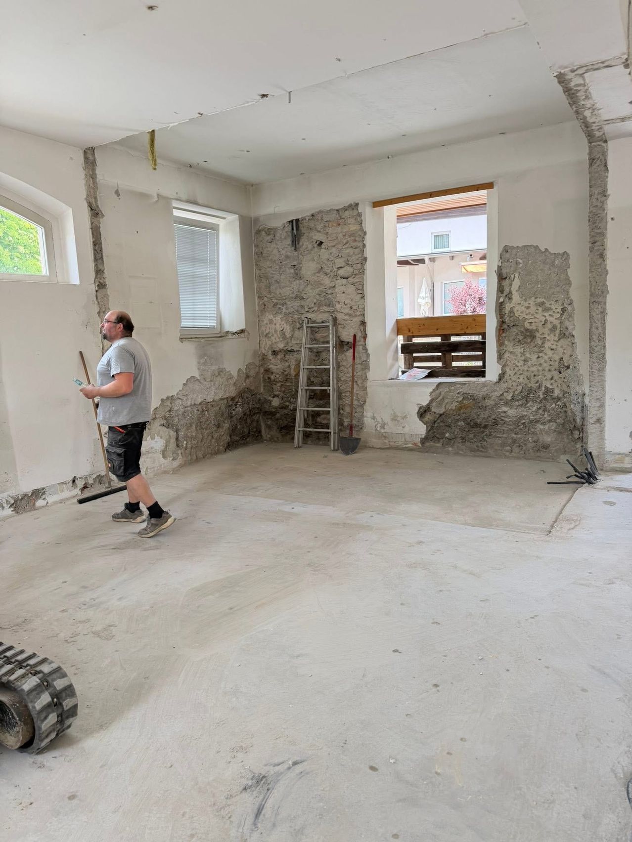 Ein Mann in einem grauen T-Shirt und Shorts arbeitet in einem Raum im Bau. Er hält einen Besen. Eine Leiter und eine Schaufel stehen an der Wand. Zwei Fenster und eine Tür sind sichtbar.