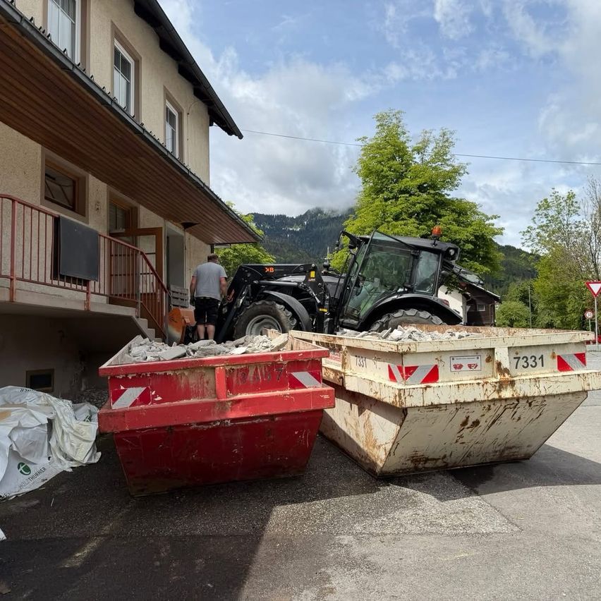 Ein Traktor mit Anhänger kippt Schutt in einen roten Müllcontainer. Ein Mann steht auf der Treppe, und ein Gebäude mit Balkon ist im Hintergrund.