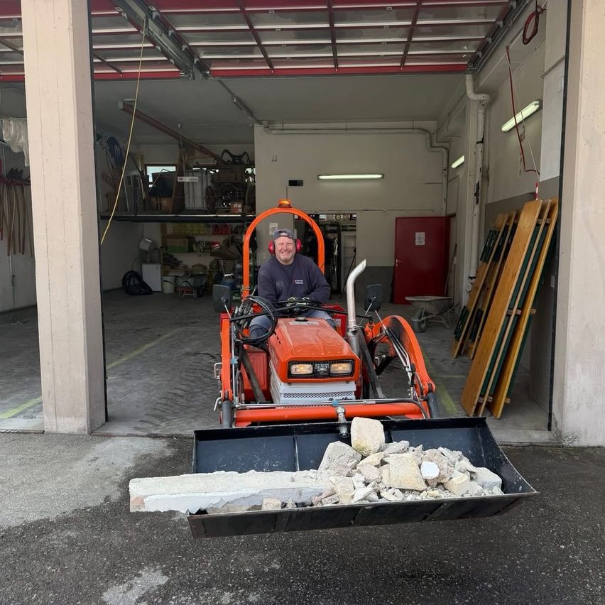 Ein Mann fährt einen orangefarbenen Traktor in einer Garage, beladen mit Schutt. Er trägt ein blaues Hemd und eine Mütze, mit einer roten Tür hinter ihm.