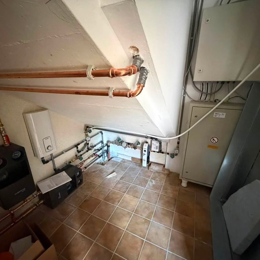 Ein Raum mit Kupferrohren und Heizgeräten. Der Boden ist gefliest, und es gibt Elektrokästen und einen Wasserboiler.